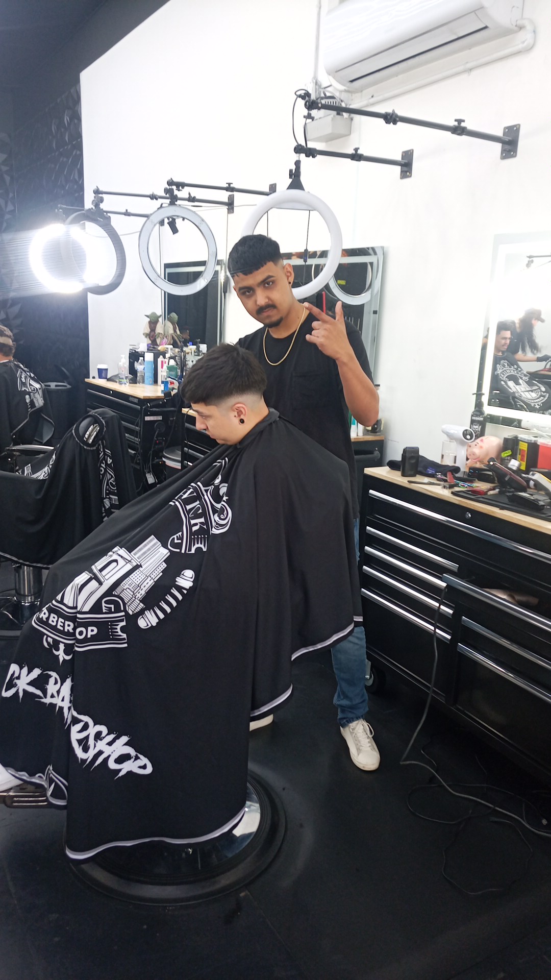 A Bushwick barber beside a client mid cut, wearing the shop's branded cape
